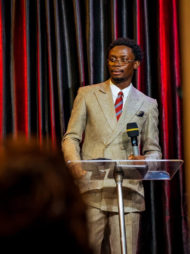 Precious Alo teaching from a clear lectern with a tablet and microphone
