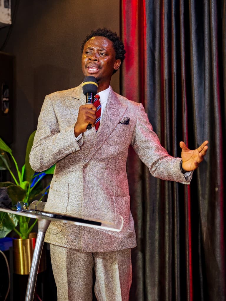 Precious Alo preaching with an open hand, microphone raised, against a burgundy-lit curtain
